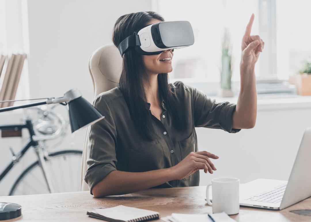 Woman in virtual reality headset sits at office desk.
