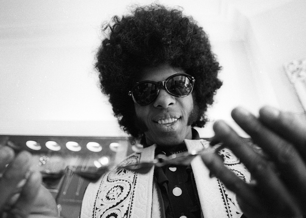 Sly Stone wearing sunglasses, holding a necklace.