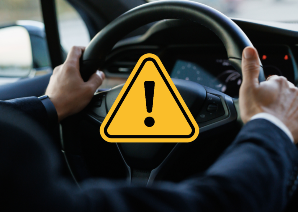 Person’s hands on steering wheel of a Tesla with yellow caution symbol overlaid in the center of the photo.