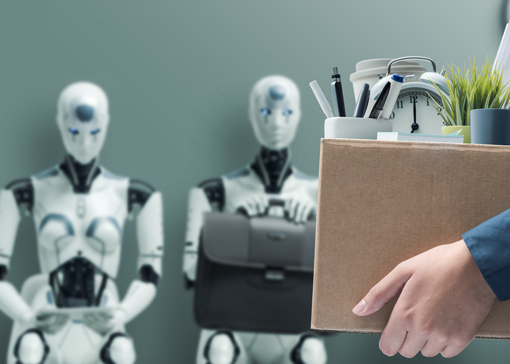 Close up of hands holding a cardboard box full of office supplies with two robots in the background appearing to look like they are waiting for a job interview.