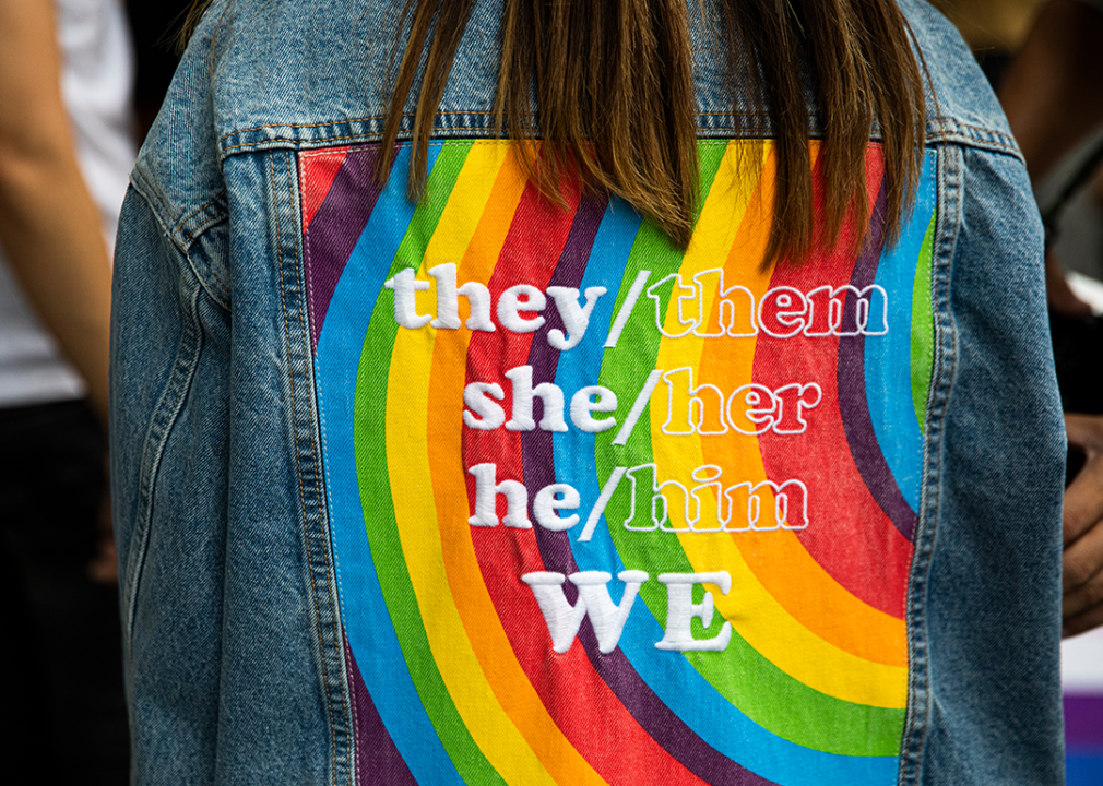 View of the back of a person wearing a denim jacket with gender neutral pronouns over a rainbow print.