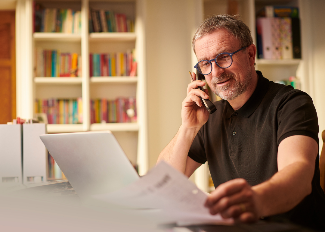 Person speaking on phone with laptop reviewing paper documents.