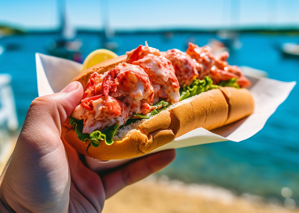 Hand holding lobster roll with summer harbor in background.