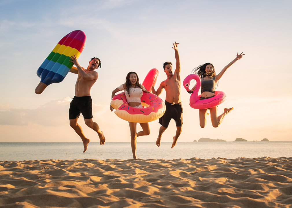 Four friends jumping on the beach holding colorful inflatable pool toys.