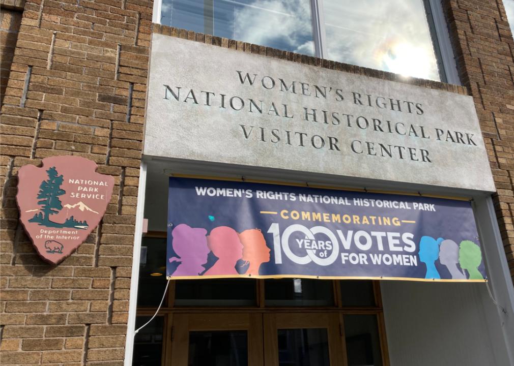A photo of the sign above the door of the Women's Rights National Historical Park Visitor Center with a banner that reads "Women's Rights National Historic Park, commemorating 100 years of votes for women.