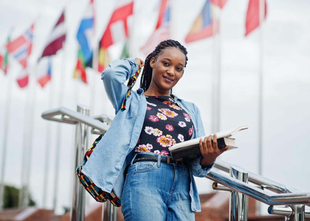 Student holding books on university campus with flags in background.