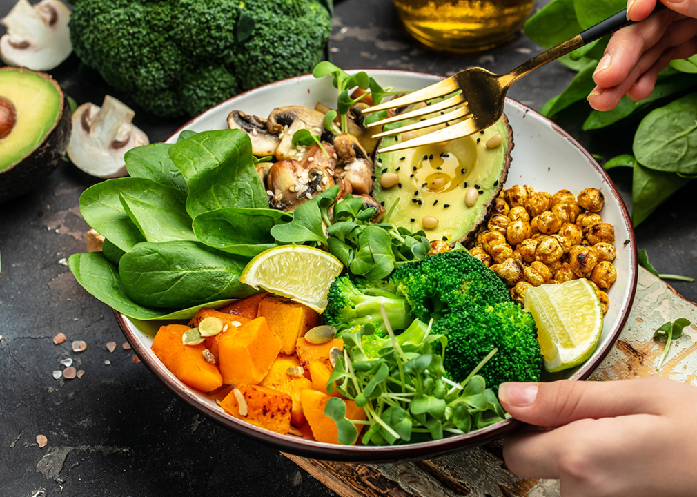 Hands holding healthy vegan bowl with spinach, broccoli, chickpeas, avocado, pine nuts, mushrooms, and squash.