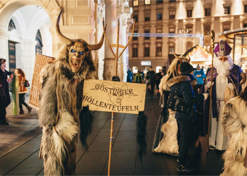 A person dressed as a scary looking Krampus walking down the street holding a sign that reads Gostinger Hollenteufeln.
