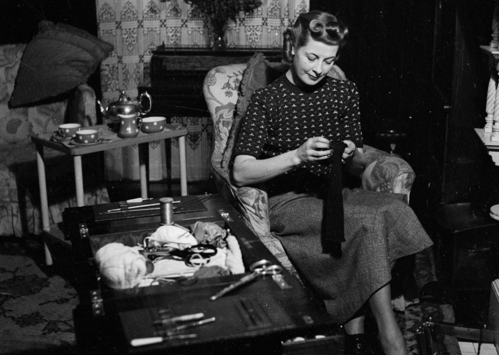 A woman sits by the fireside darning socks, with an open sewing table by her side.