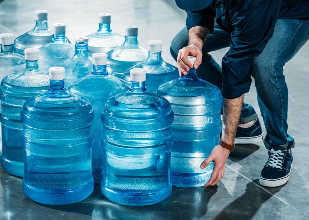 Photo shows a dozen water jugs and a person bending down to pick one up