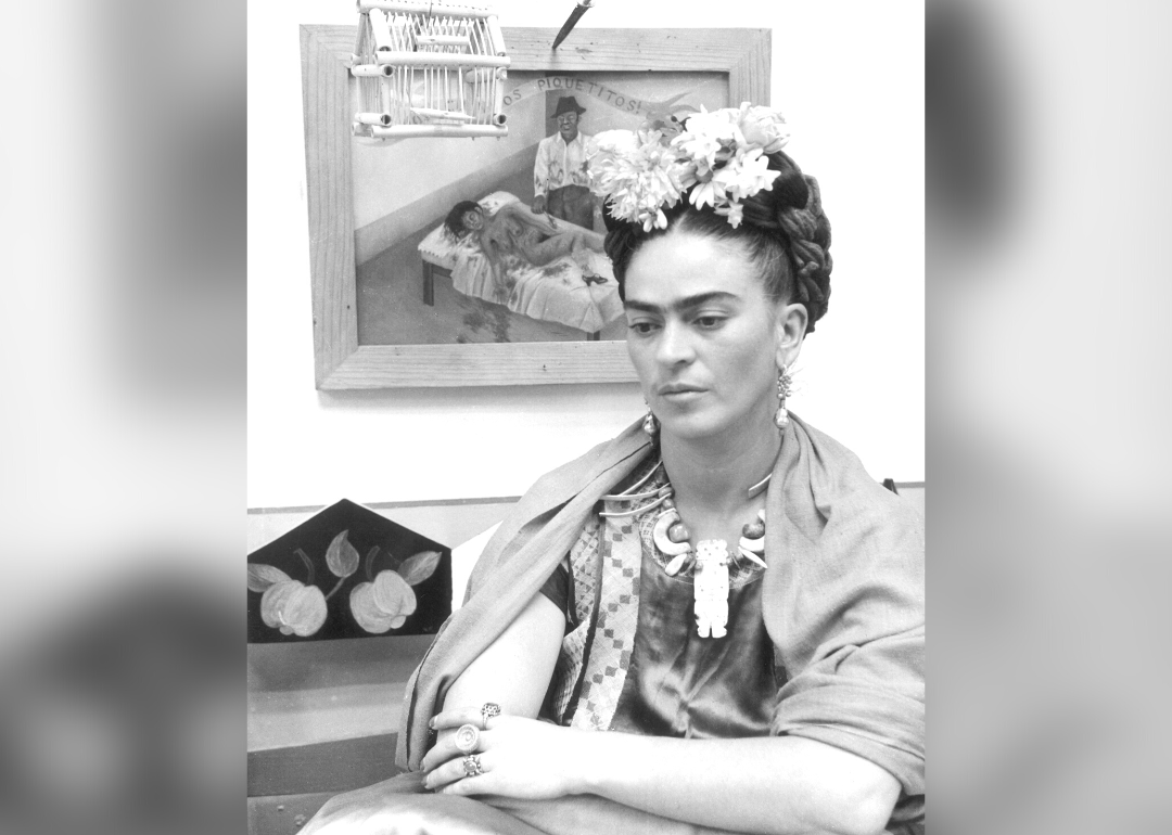 Frida Kahlo seated with arms folded in front of one of her paintings.
