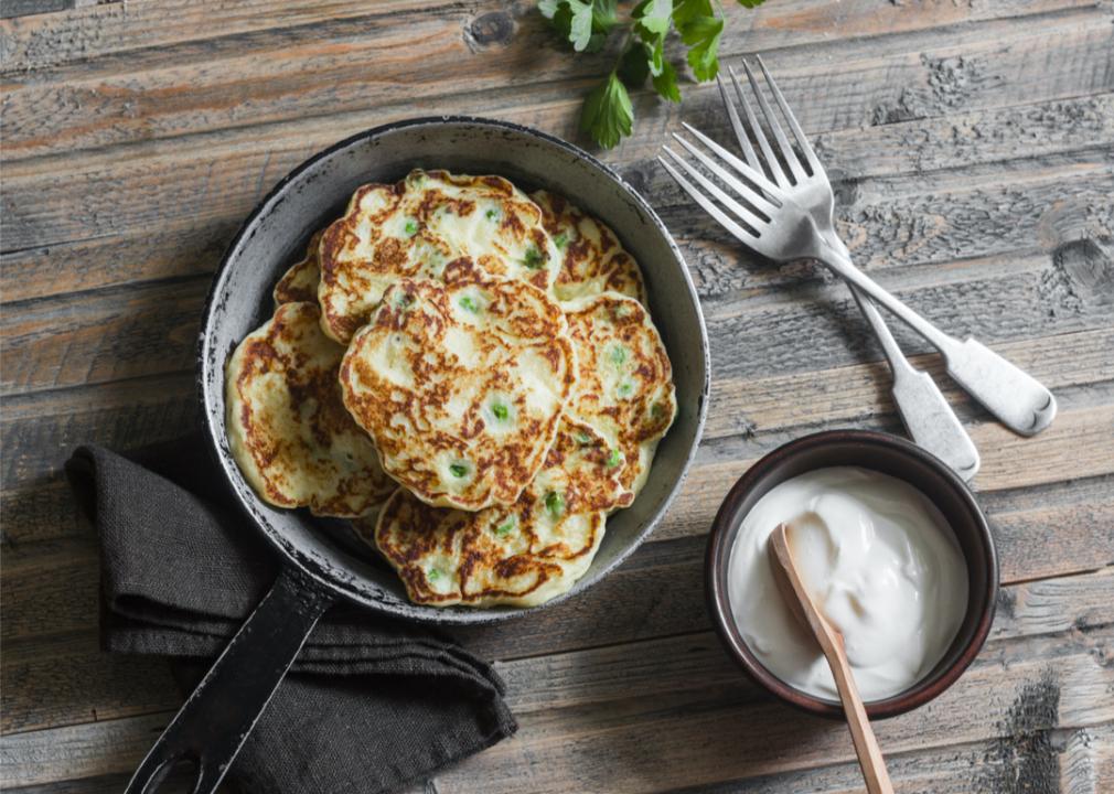 Fried leek patties in a skillet next to a bowl of sour cream.