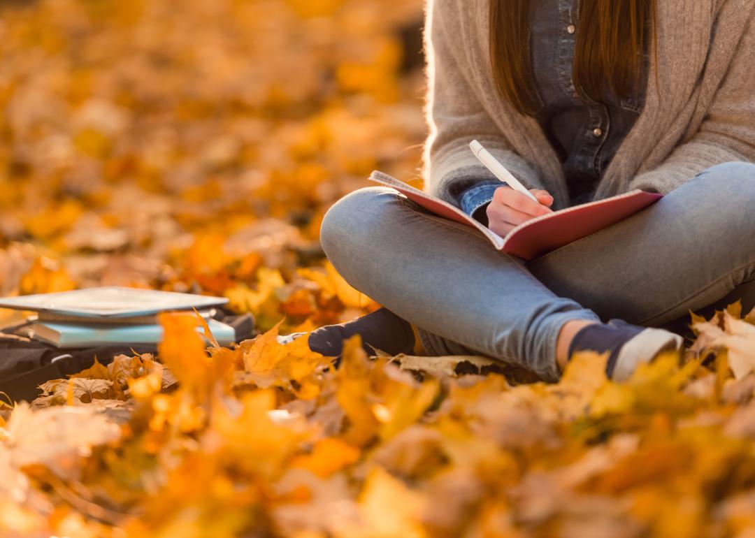 Cropped view of student sitting in autumn leaves writing in notebook.