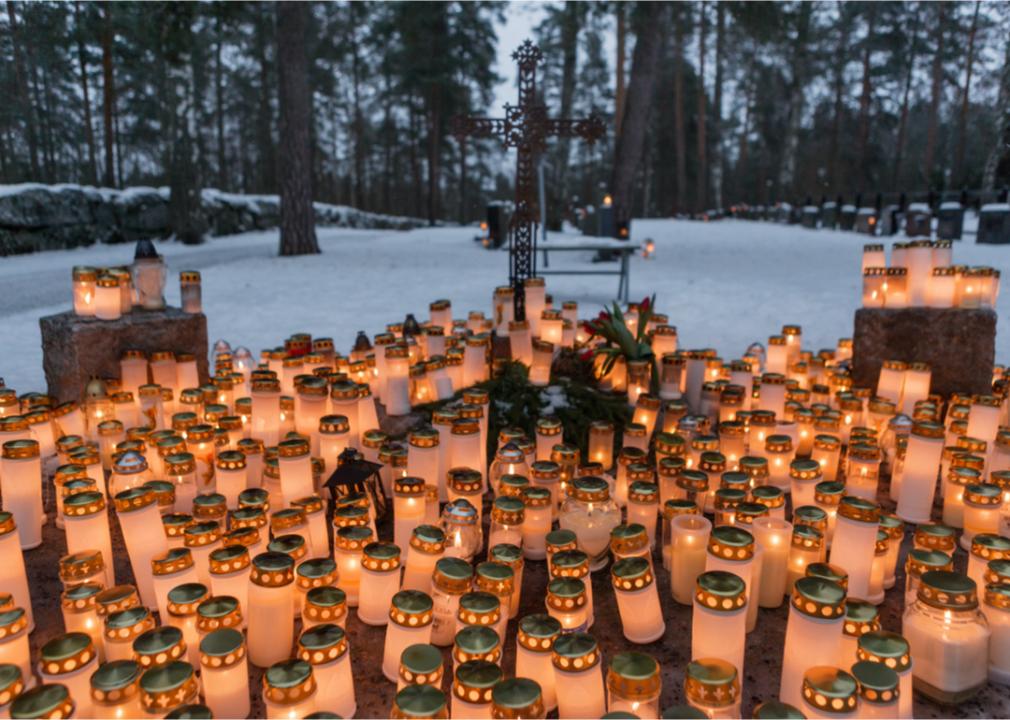 Many lit lanterns placed in front of an iron cross surrounded by snowy woods.