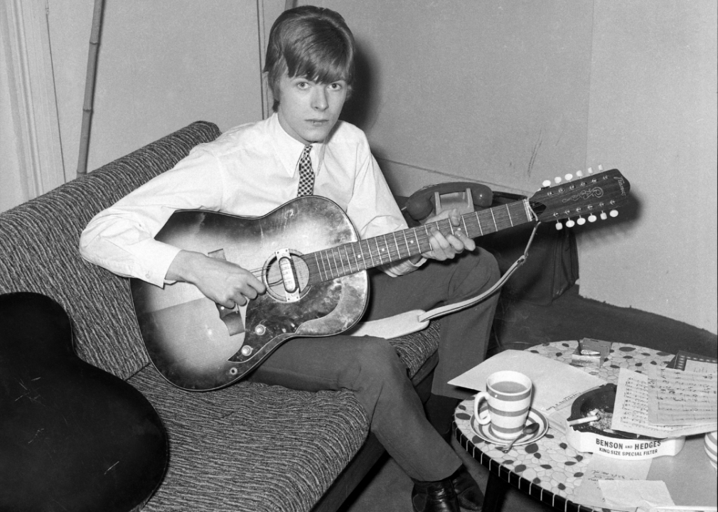 A young Bowie sitting on a couch playing the guitar.
