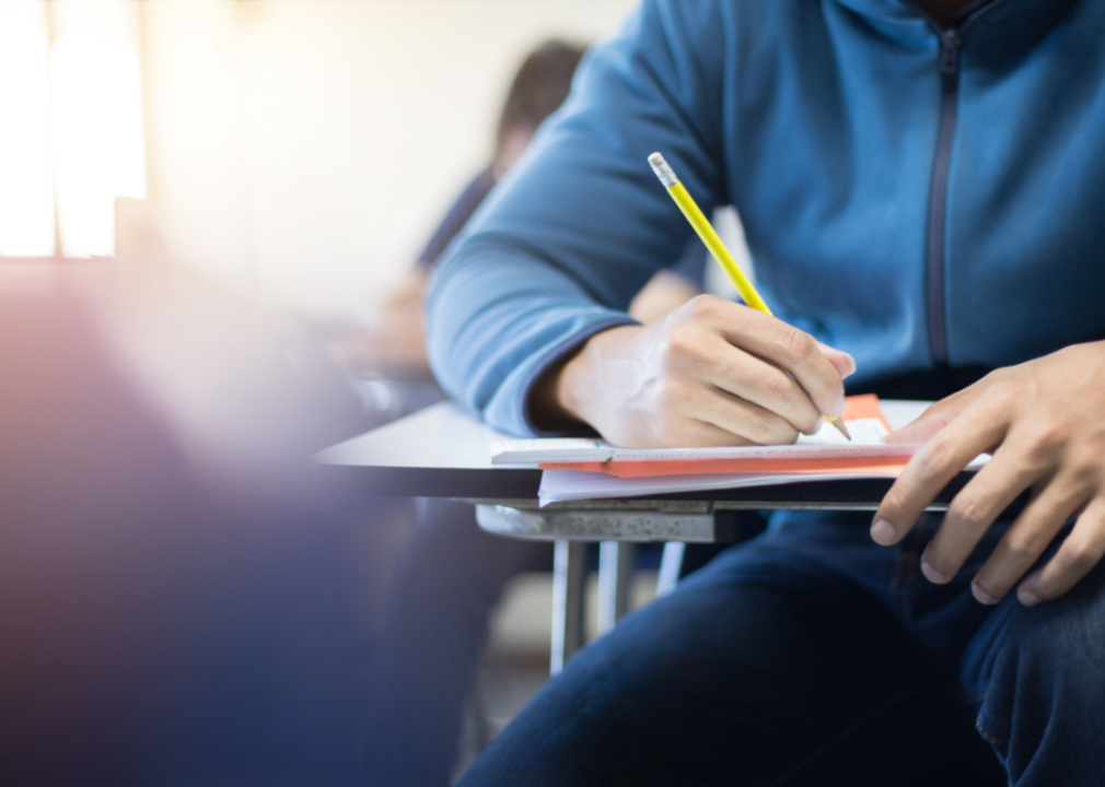 A student's hand takes notes at a desk in a classroom