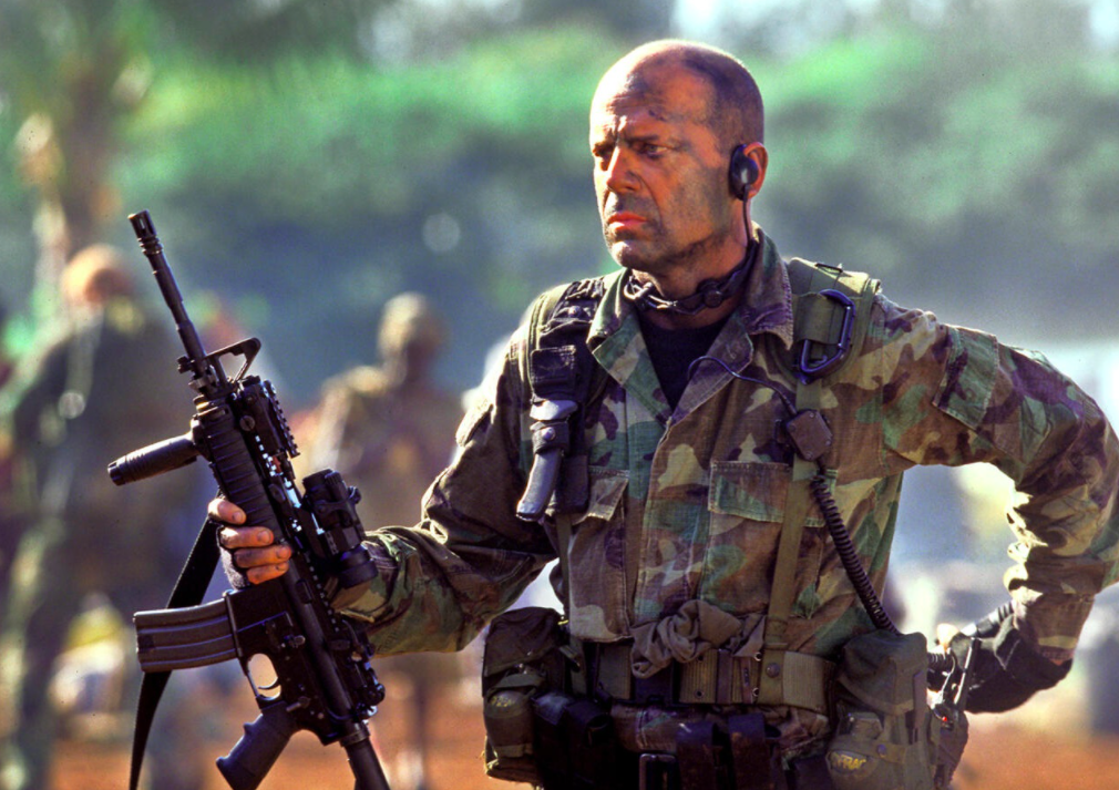 Bruce Willis in full camo and painted face holding a large gun.