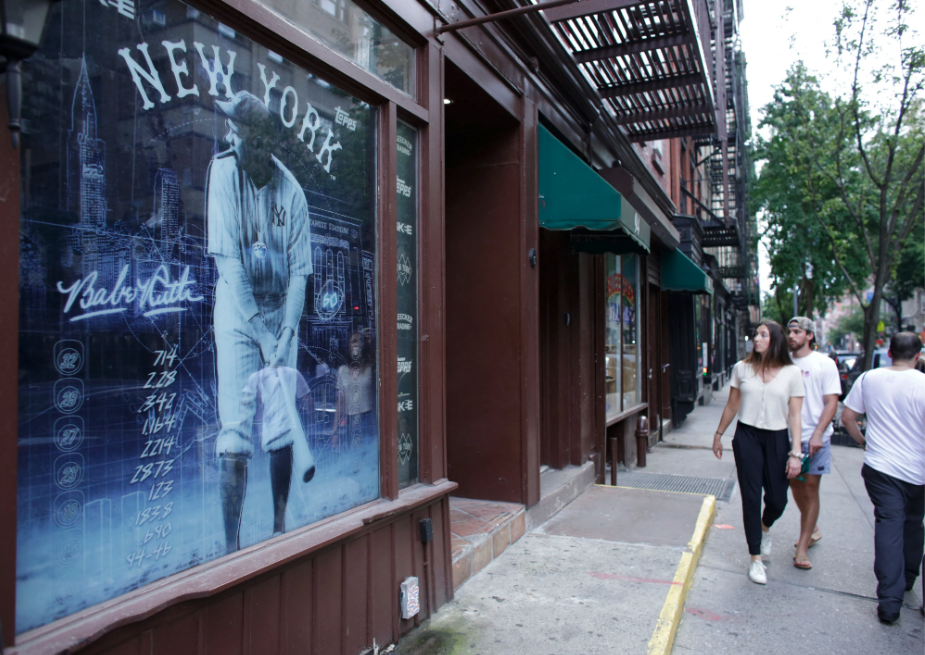 People pass by Bleecker Trading in New York on July 06, 2021 looking at a black and white image of Babe Ruth covering the window.