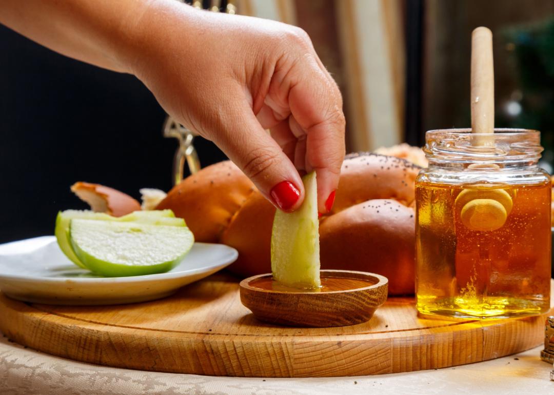 A person's hand dips a piece of apple in honey in honor of the celebration of Rosh Hashanah near honey and challah and traditional food.