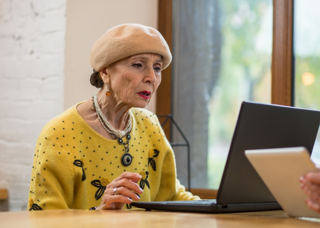An older woman in a yellow sweater reads on her laptop.