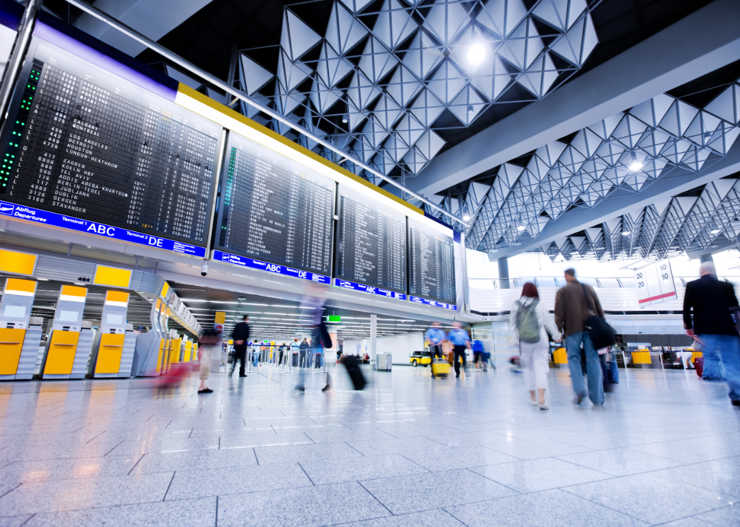 People in an airport lobby crossing in front of a plane arrival and departure board