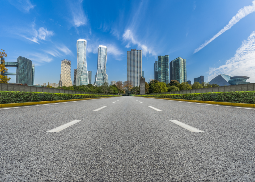 A paved road leads to a city with skyscrapers against a blue sky.