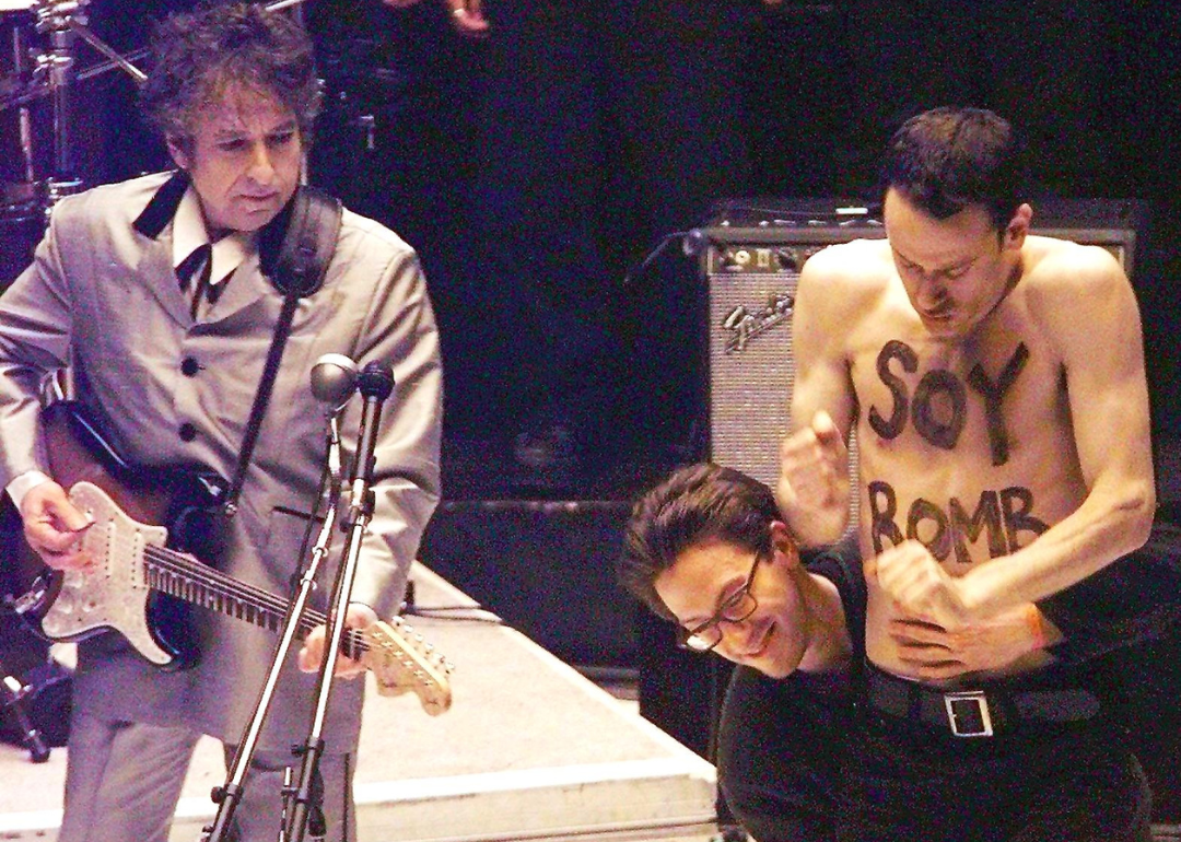 Bob Dylan performing onstage next to Michael Portnoy, who is not wearing a shirt and has writing on his chest.
