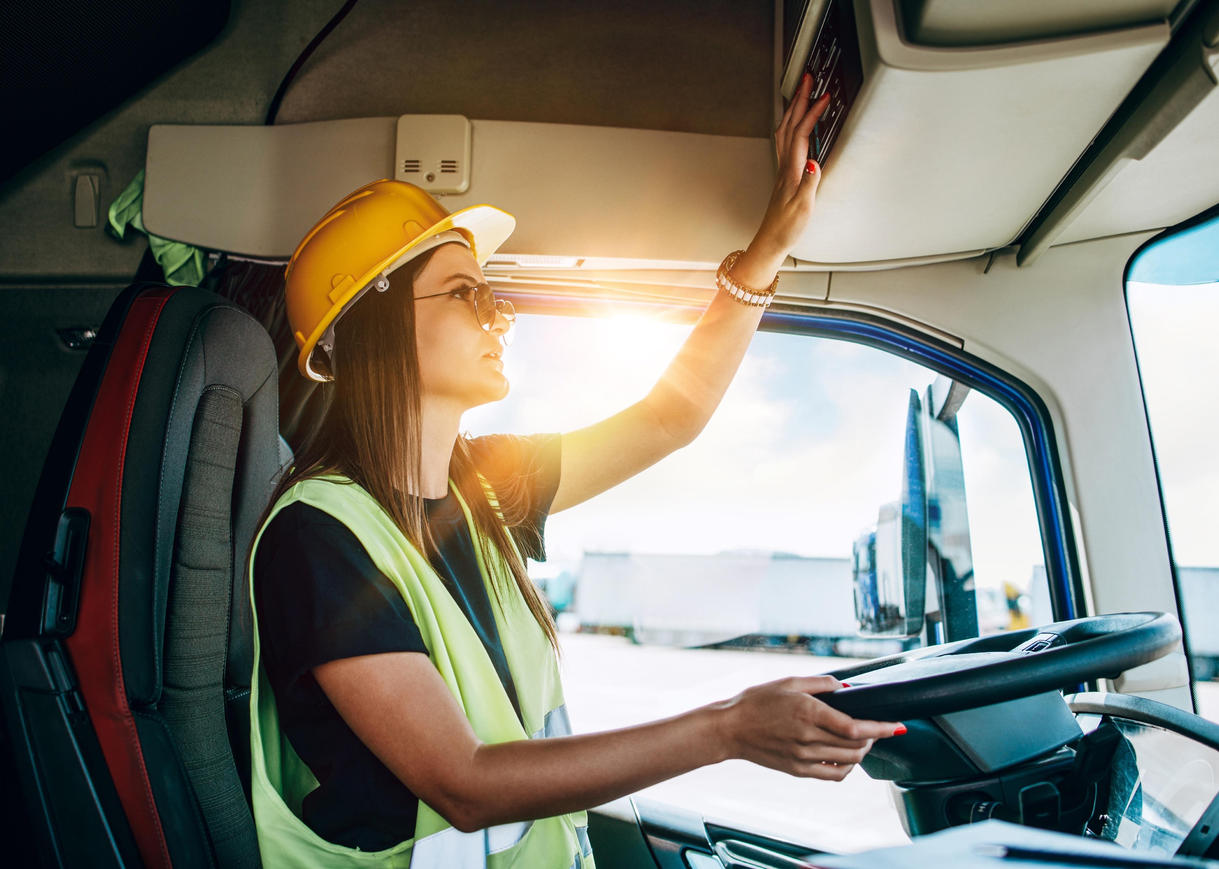 Truck driver behind the wheel wearing a hard hat and safety vest.