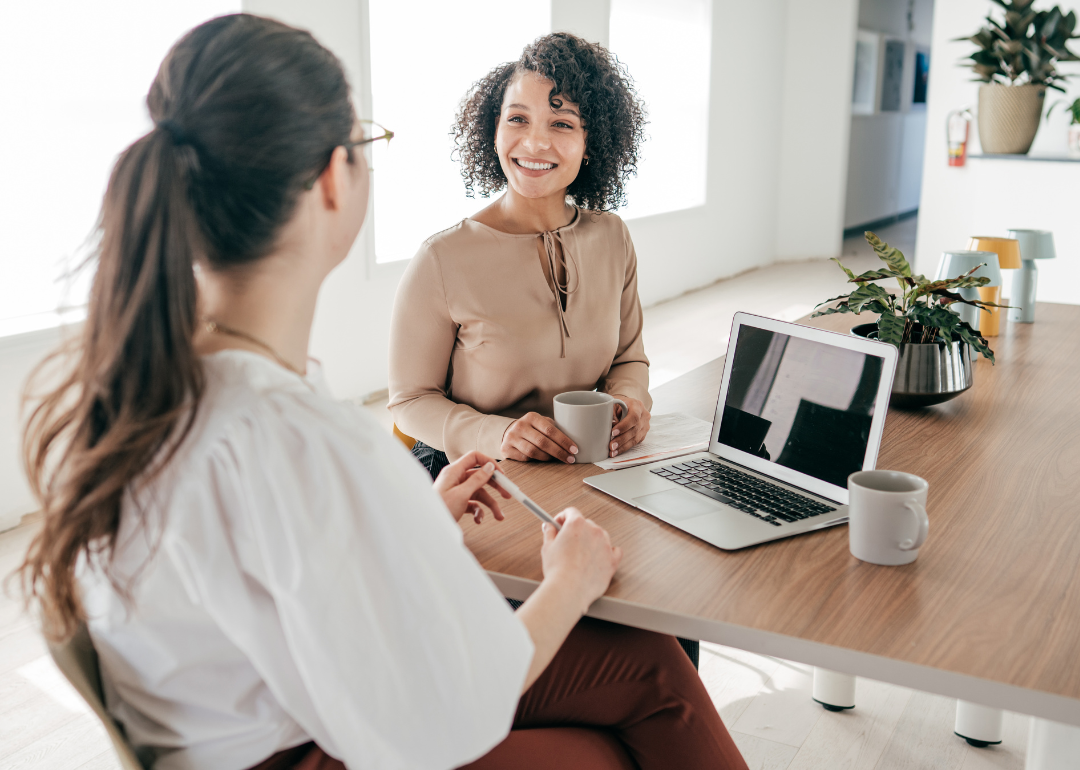 Two workers sit at a desk and talk over coffee.