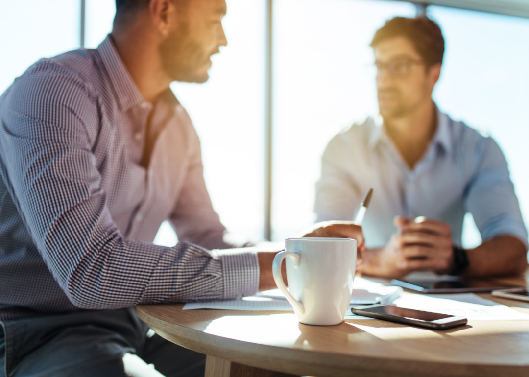 Two workers sit at a desk and talk.