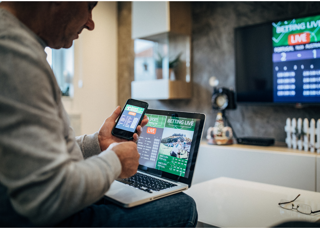 Man sitting on couch with phone, laptop, and TV on sports betting screens.