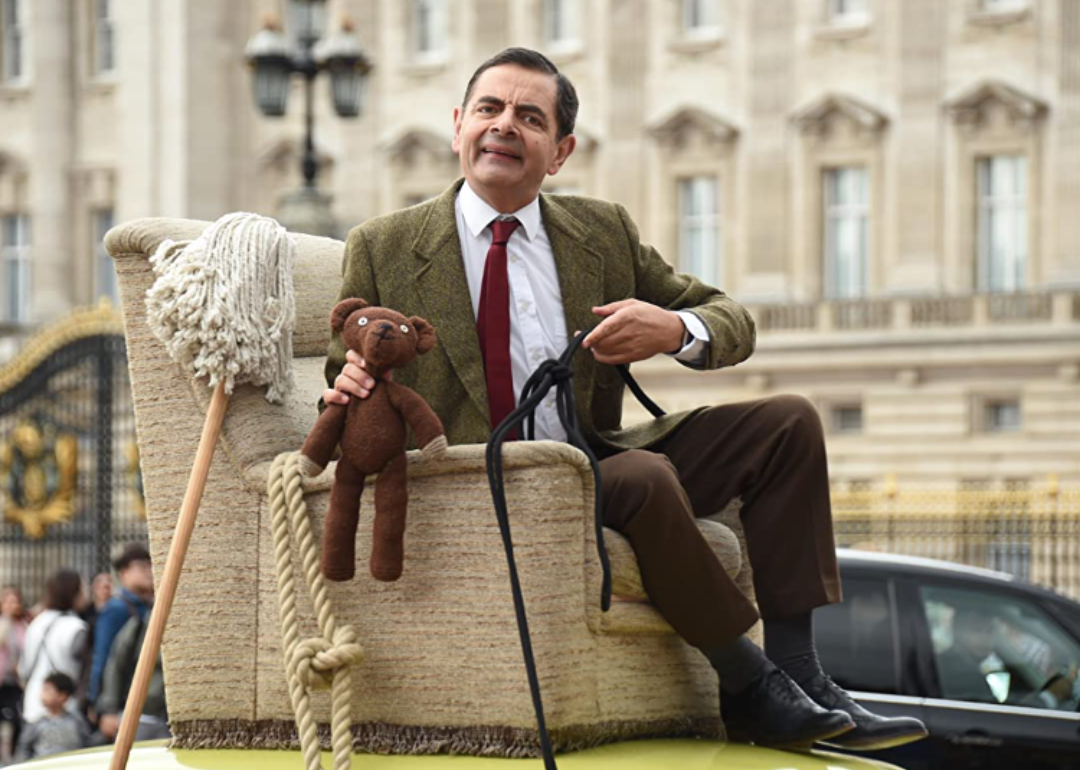 Rowan Atkinson as Mr. Bean sitting on an armchair strapped to the top of a car. 