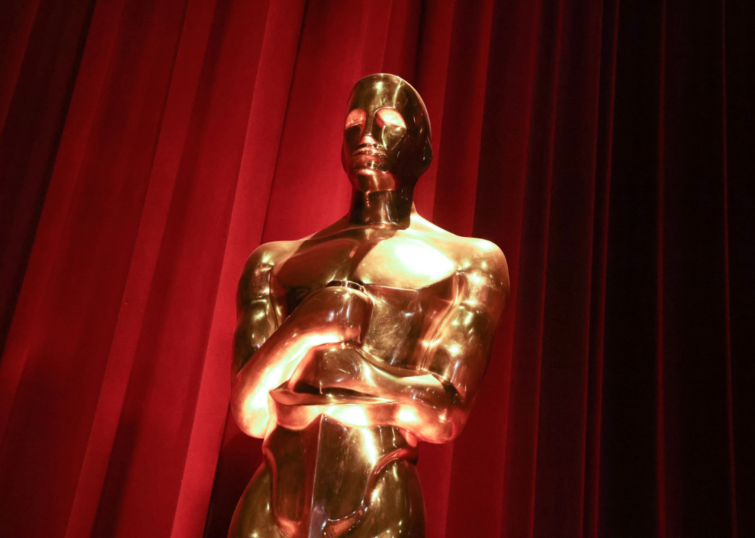 A golden Oscar statue reflects the light in front of a red velvet curtain.
