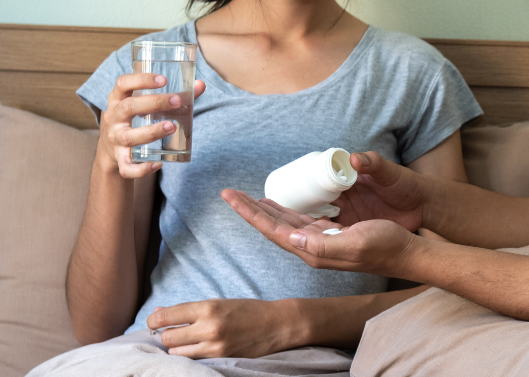Photo shows a closeup of someone pouring pills out of a pill bottle and another person sitting nearby holding a glass of water