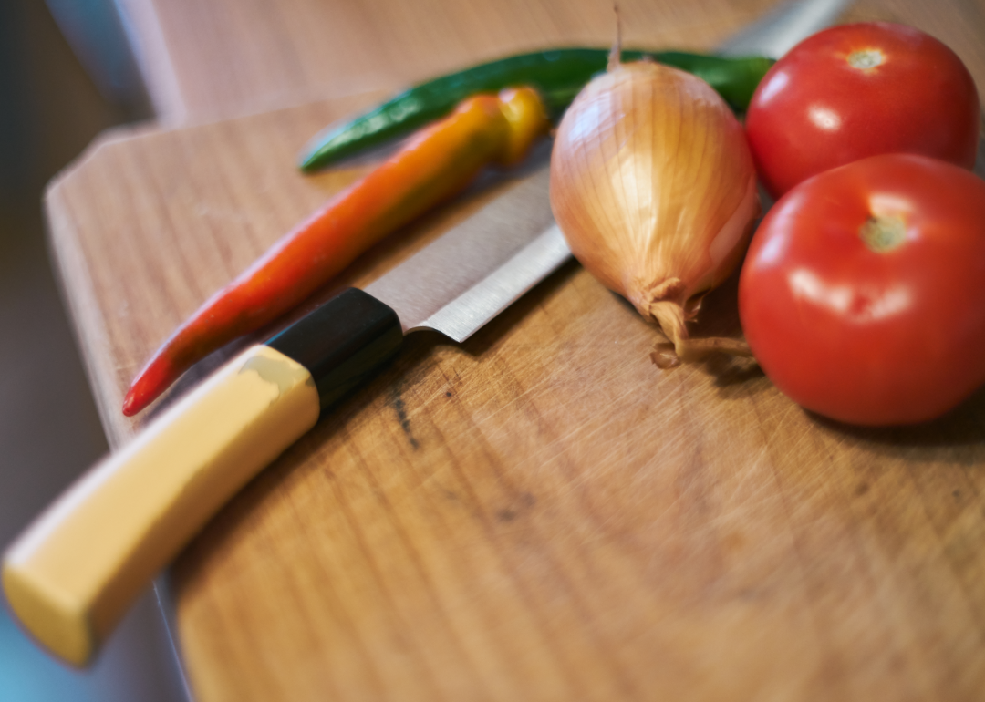 A kitchen knife rests atop a cutting board with several vegetables.