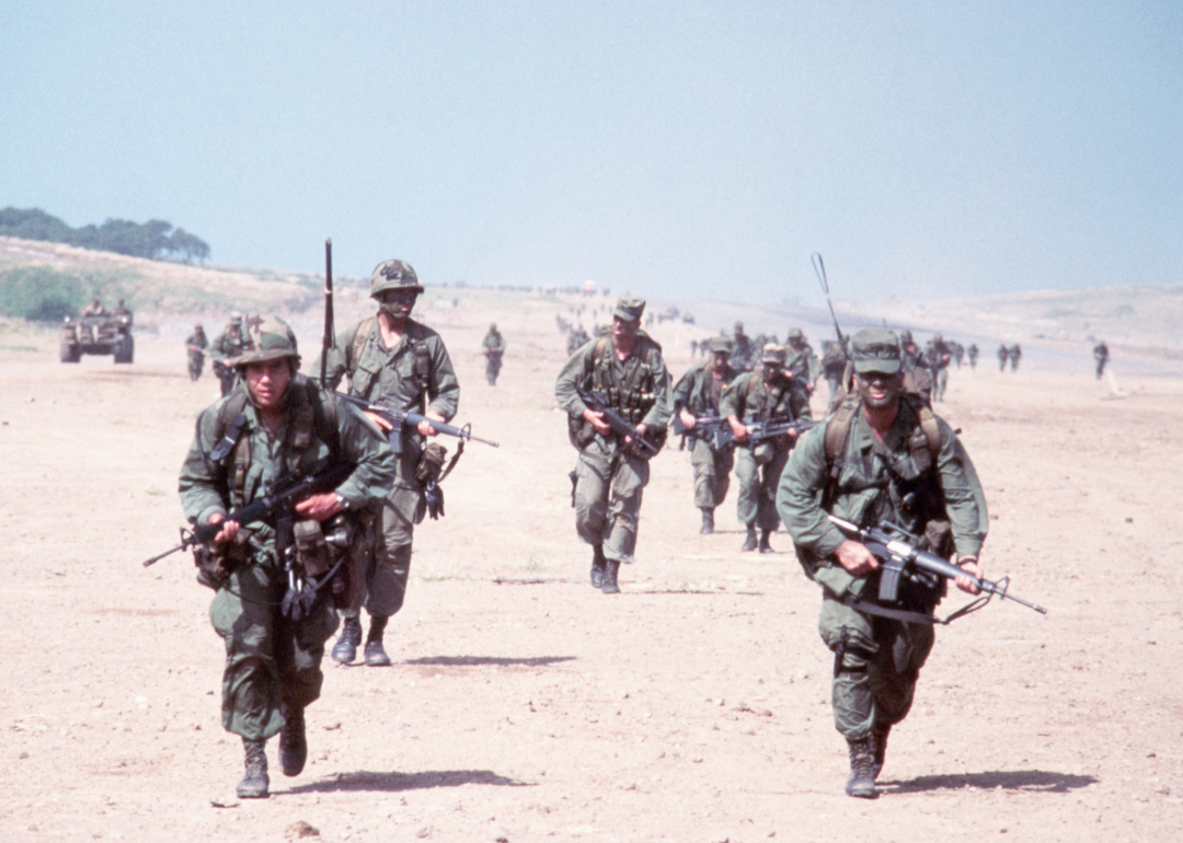 United States combat soldiers run in parallel lines across an empty dirt field during the invasion of the island of Grenada.