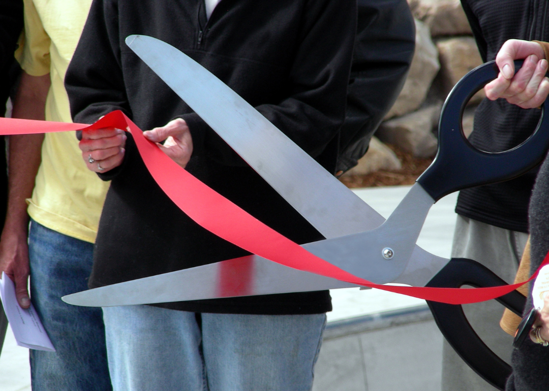 A person holds a pair of oversized scissors as they cut a red ribbon.