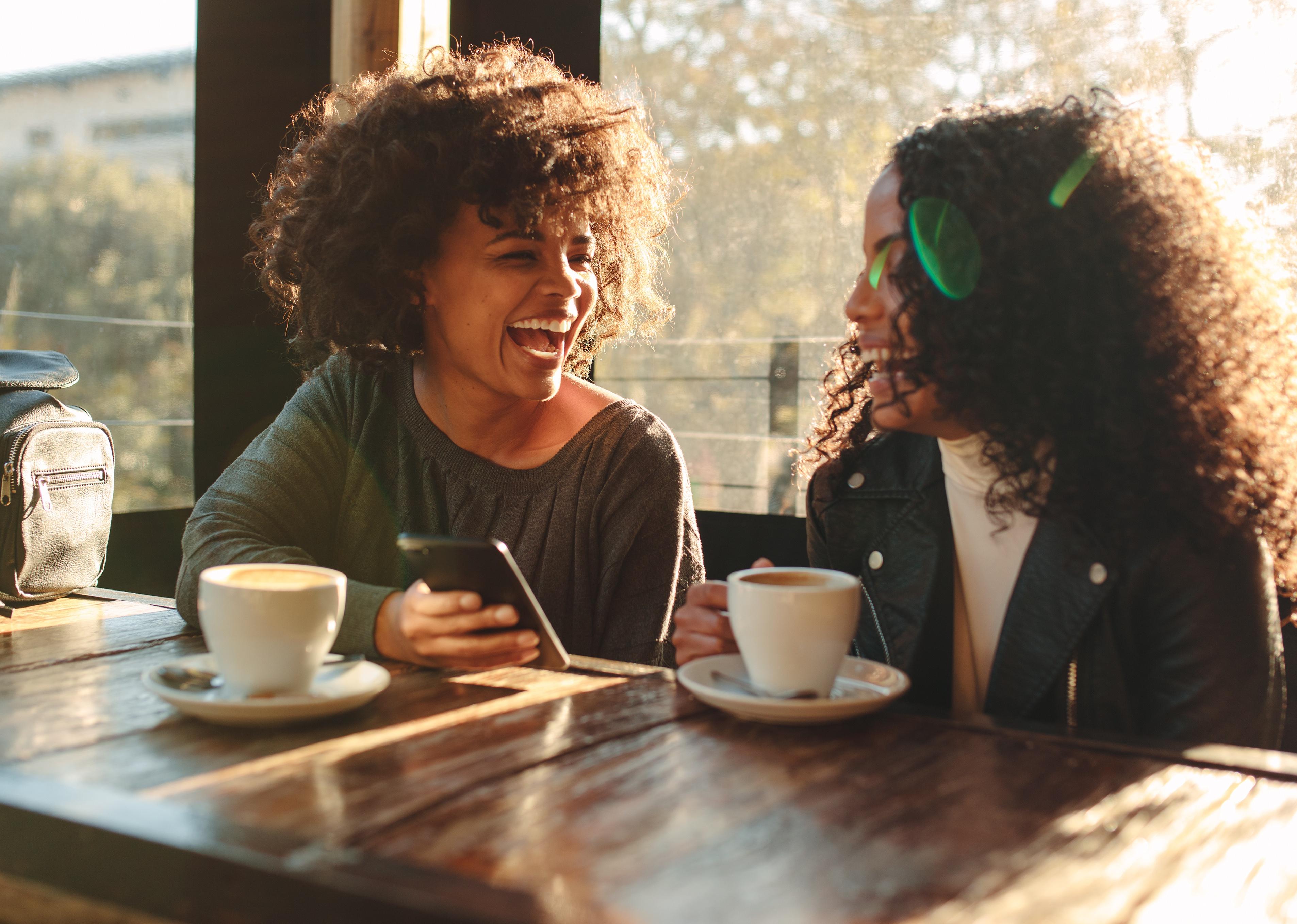 Two women laughing looking at a mobile phone inside a coffee shop.