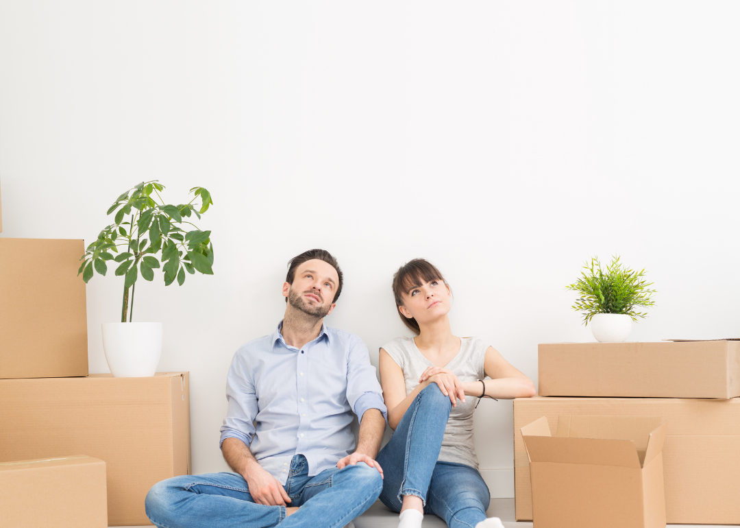 A young couple sits together amidst cardboard packing boxes preparing to move into a new home