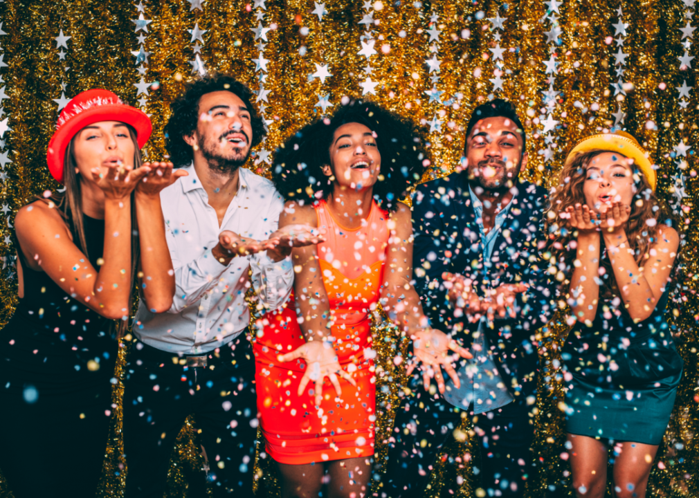 Five party guests in colorful outfits blow glitter confetti into the air.