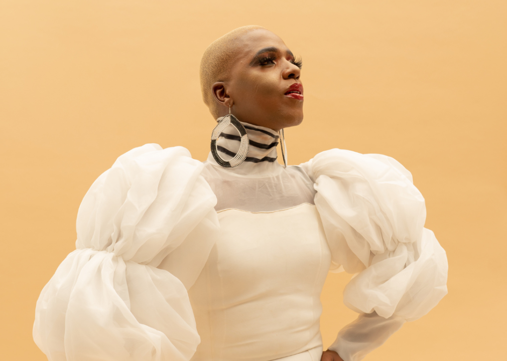 Black drag queen wearing fluffy white dress in front of yellow background.