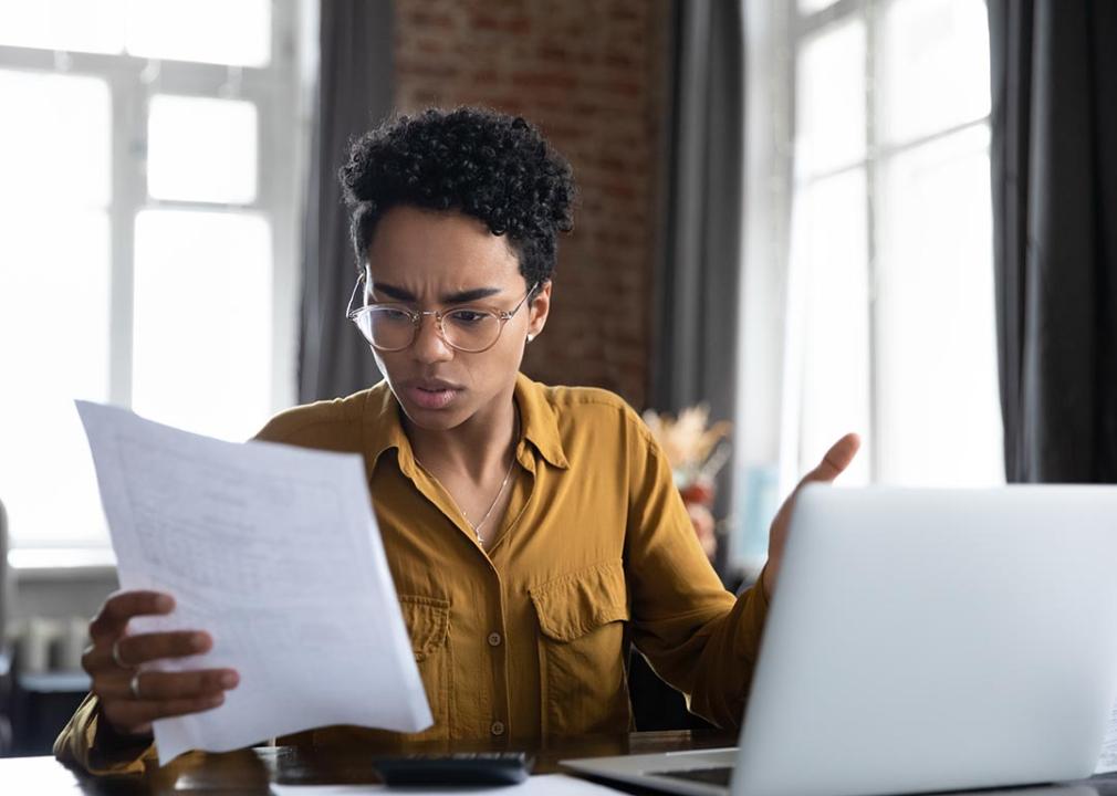 young person looks irritated while looking over paperwork in front of laptop with windows in background