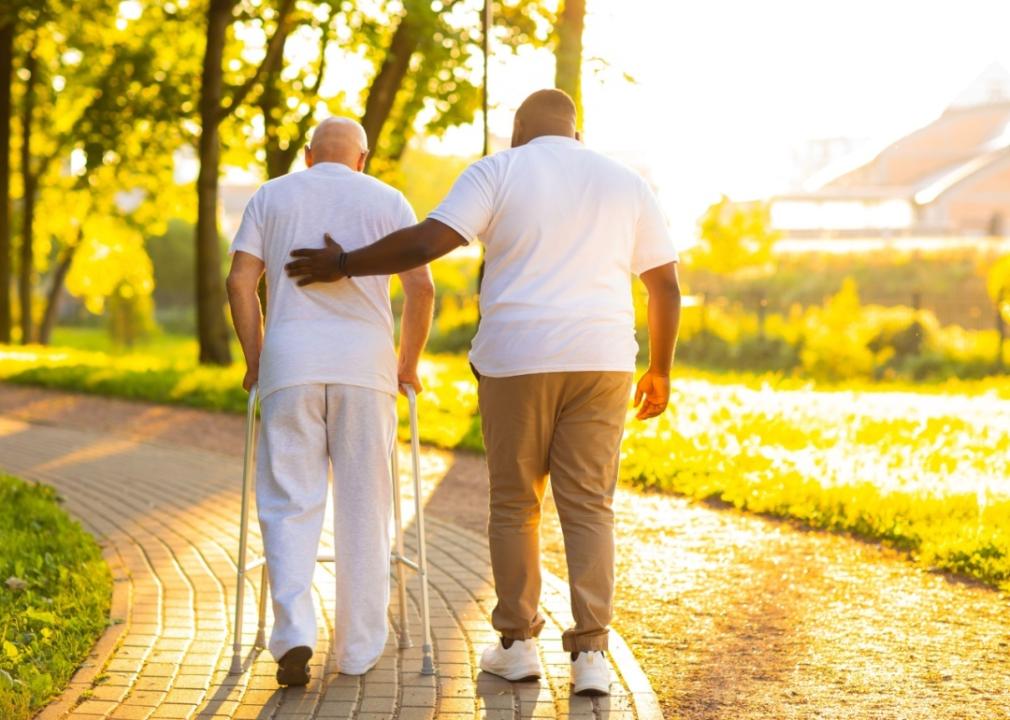 A man in a white shirt helping an elderly man use a walker in a park.