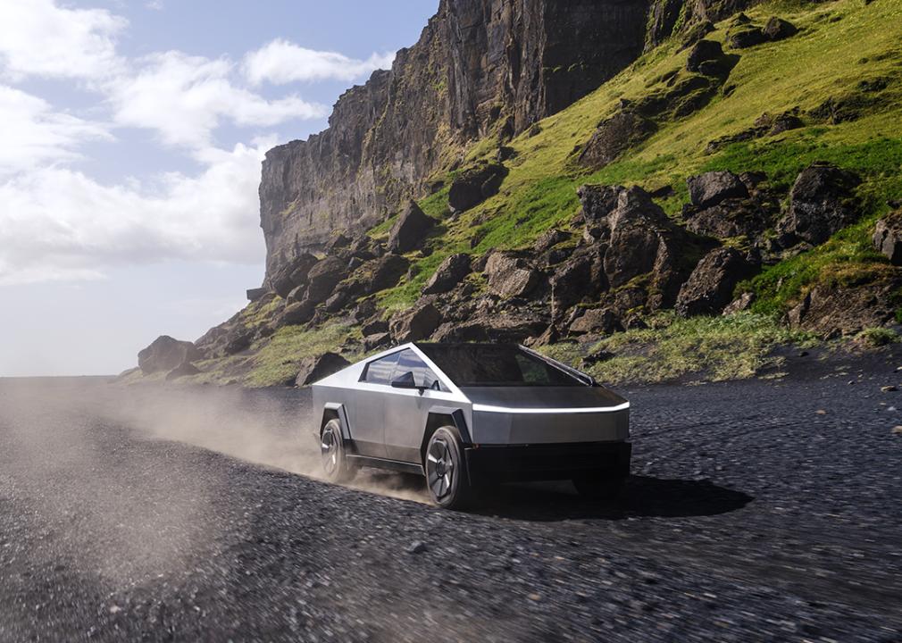 Tesla cybertruck driving on rocky beach with cliffs in background.