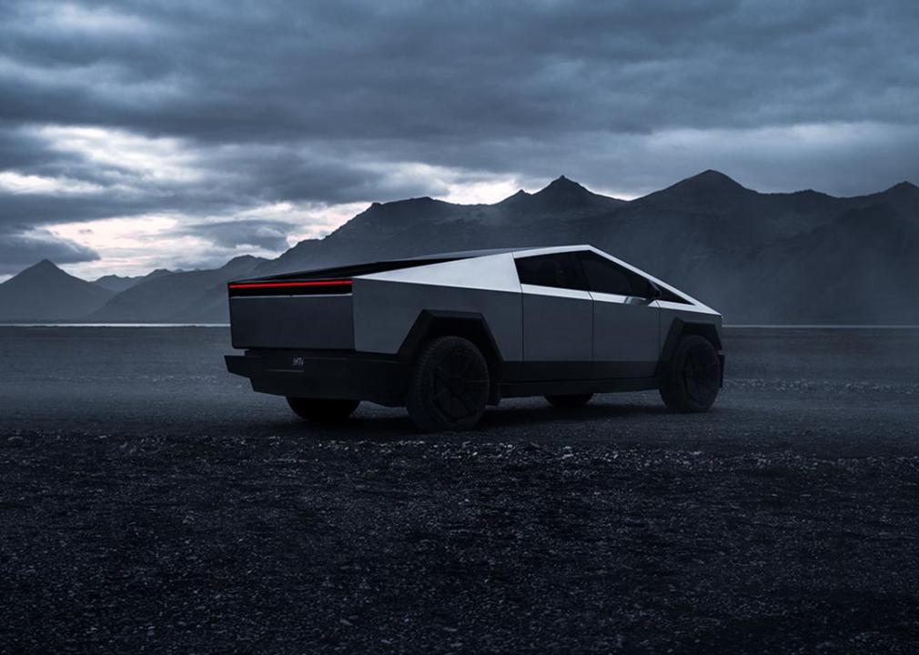 Rear 3/4 view of Tesla Cybertruck against a gray sky, dark road and mountains in background. 