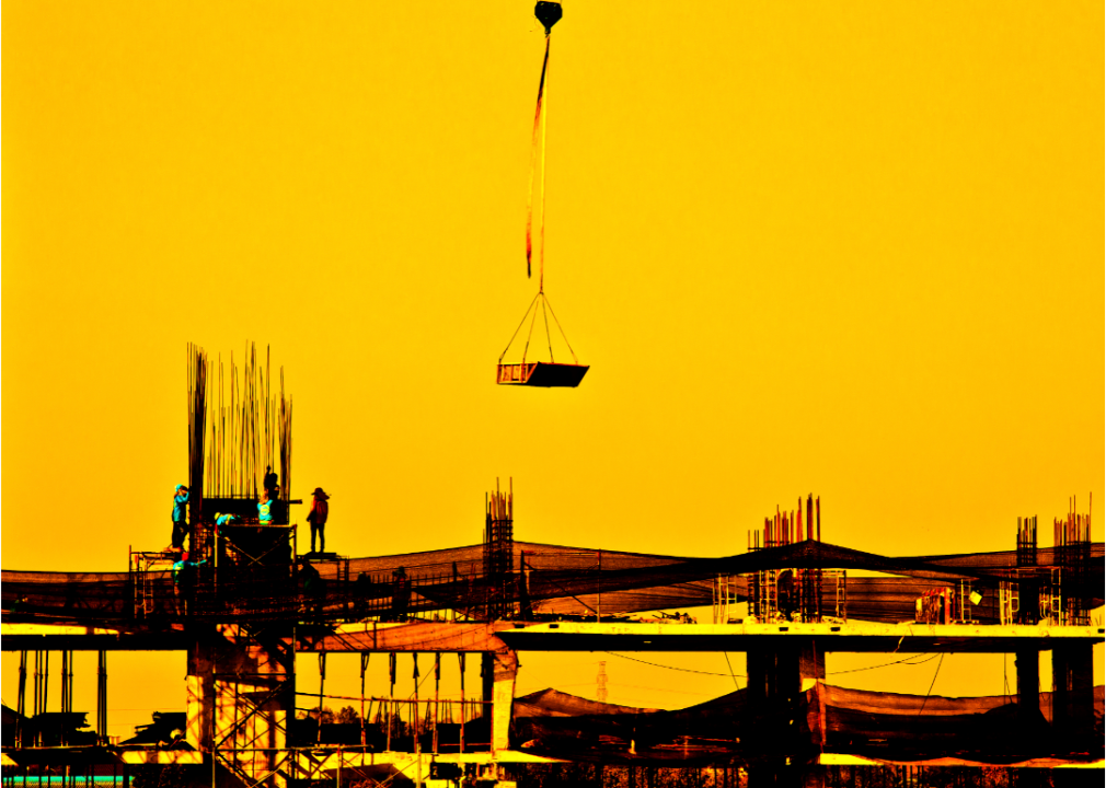 Silhouetted construction workers standing on girders wait for a crane lowering cargo with sunset yellow in the background.
