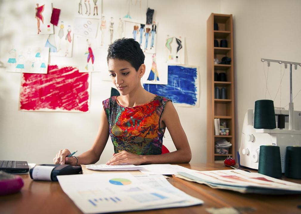 Business owner reviewing costs and paperwork sitting at desk with sewing machine and drawings visible behind her.