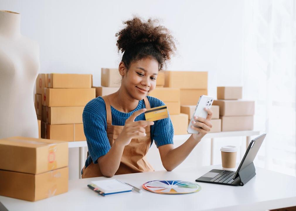 Woman holds credit card with one hand while reviewing purchases in small business setting with packages behind her. 