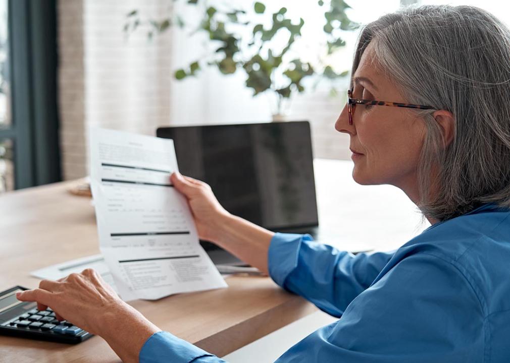A middle-aged woman sitting with a laptop and holding a bill while entering numbers in a calculator.