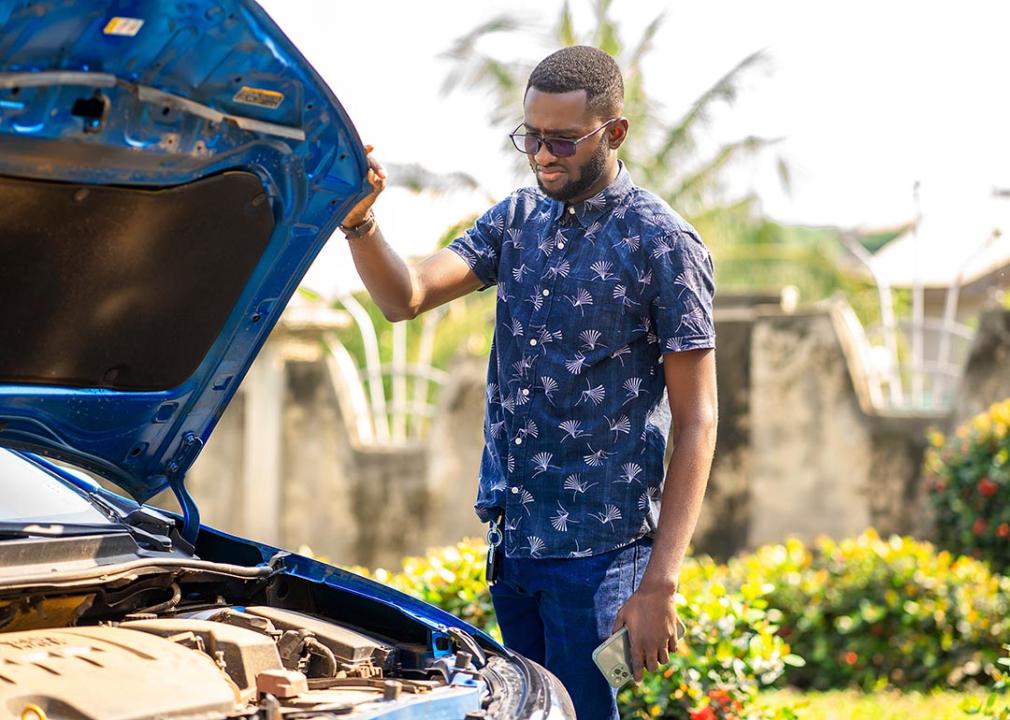 Man with sunglasses stands with the hood of a blue car open, looking at engine, with a nice yard in the background.